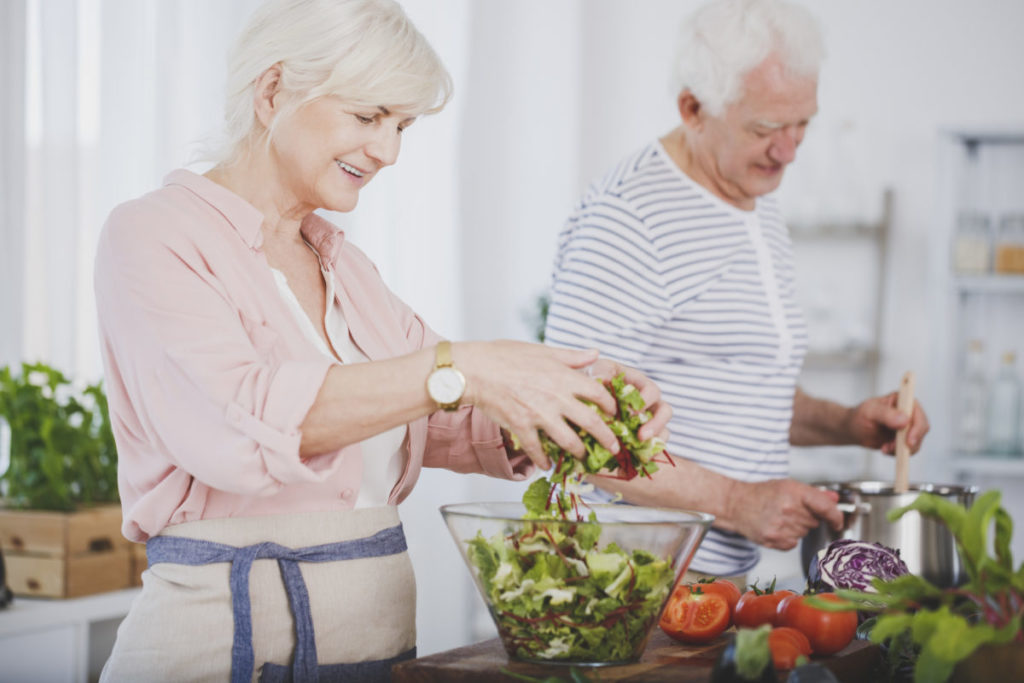 Older woman tossing a salad and her husband stirring food in a pot, cooking together 